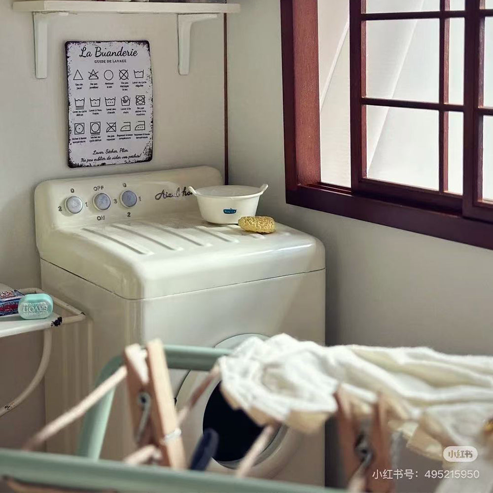 Off-white miniature washing machine beneath a laundry sign beside a window, with doll clothes on a drying rack in the foreground