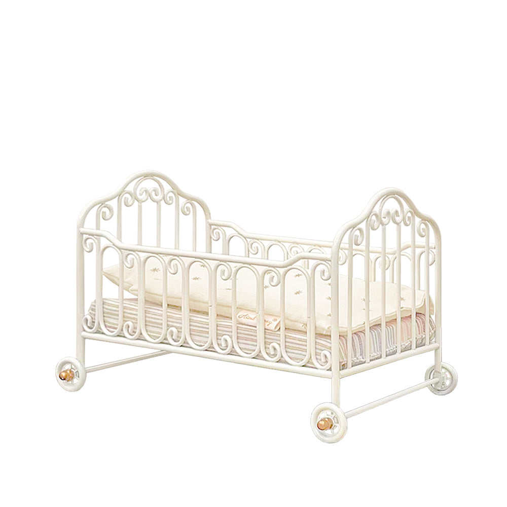 Cream-white metal miniature baby cot without canopy or support, with scrollwork rails, rolling wheels and cotton pillow, quilt and striped mattress on a white background