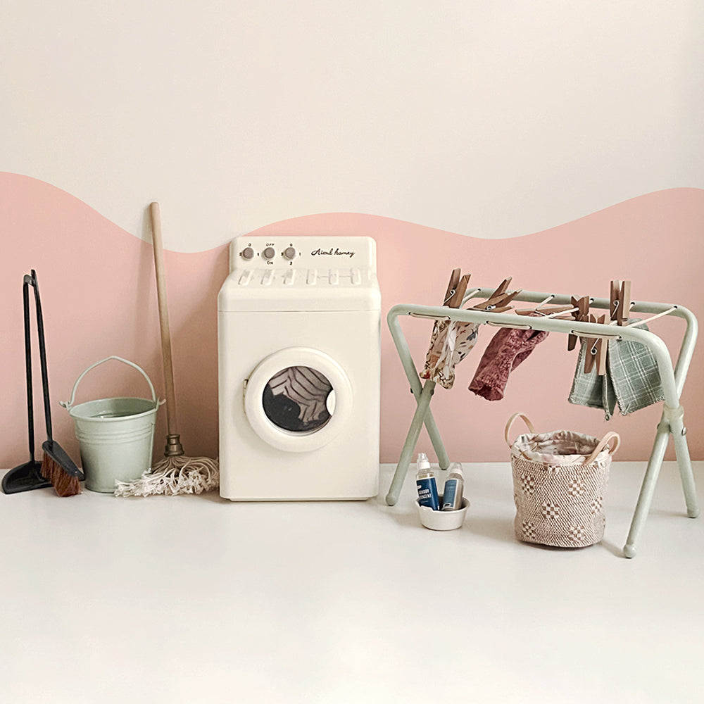 Off-white miniature washing machine styled beside a mint drying rack with doll clothes, bucket, broom, mop and laundry basket